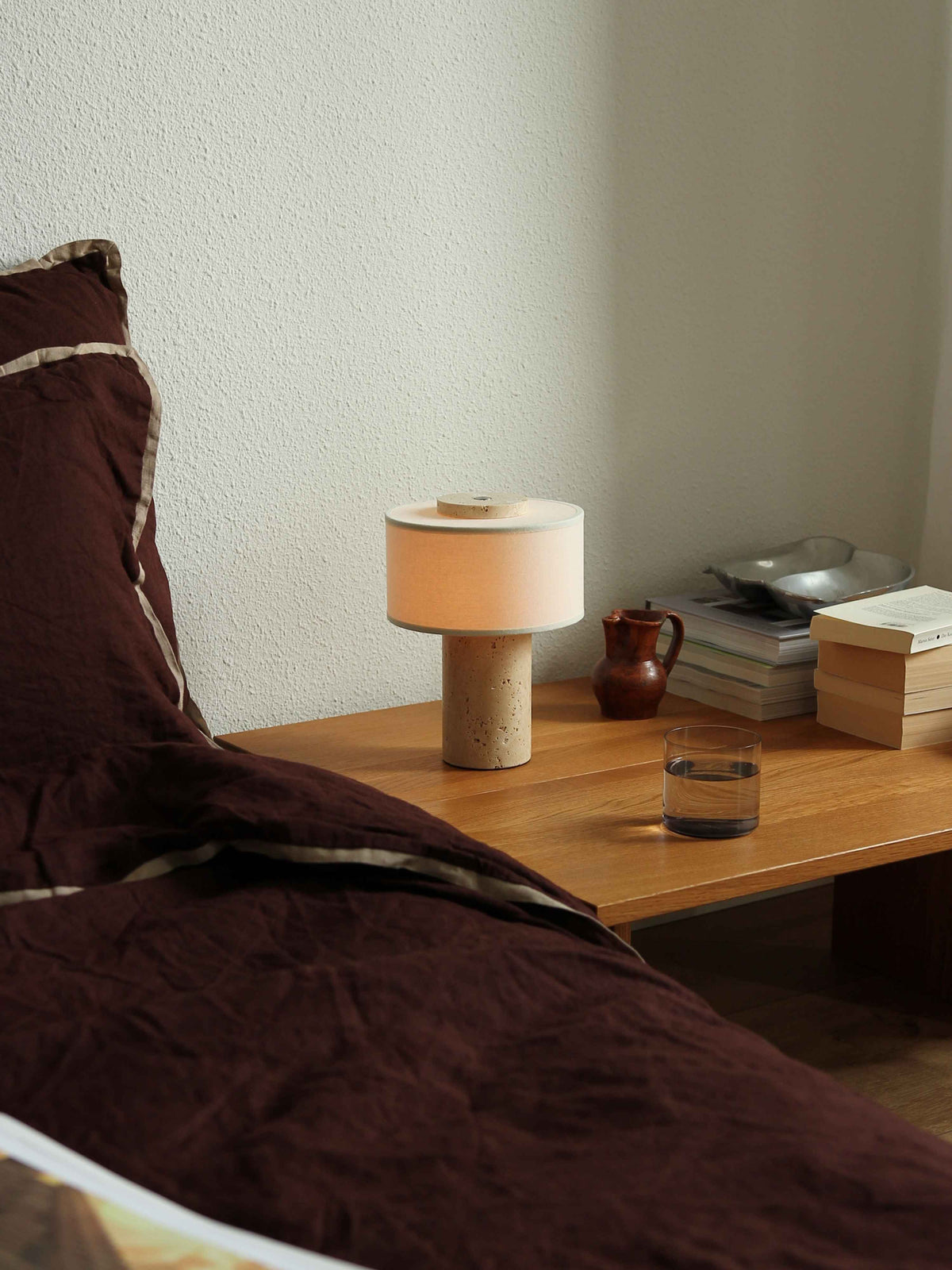Oriel Travertine Portable Lamp on a bedside table beside dark bedding